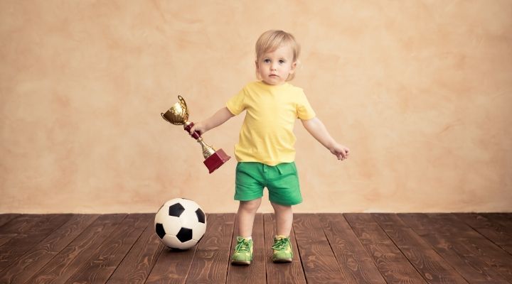 A toddler holding a trophy while a soccer ball is on the floor beside her
